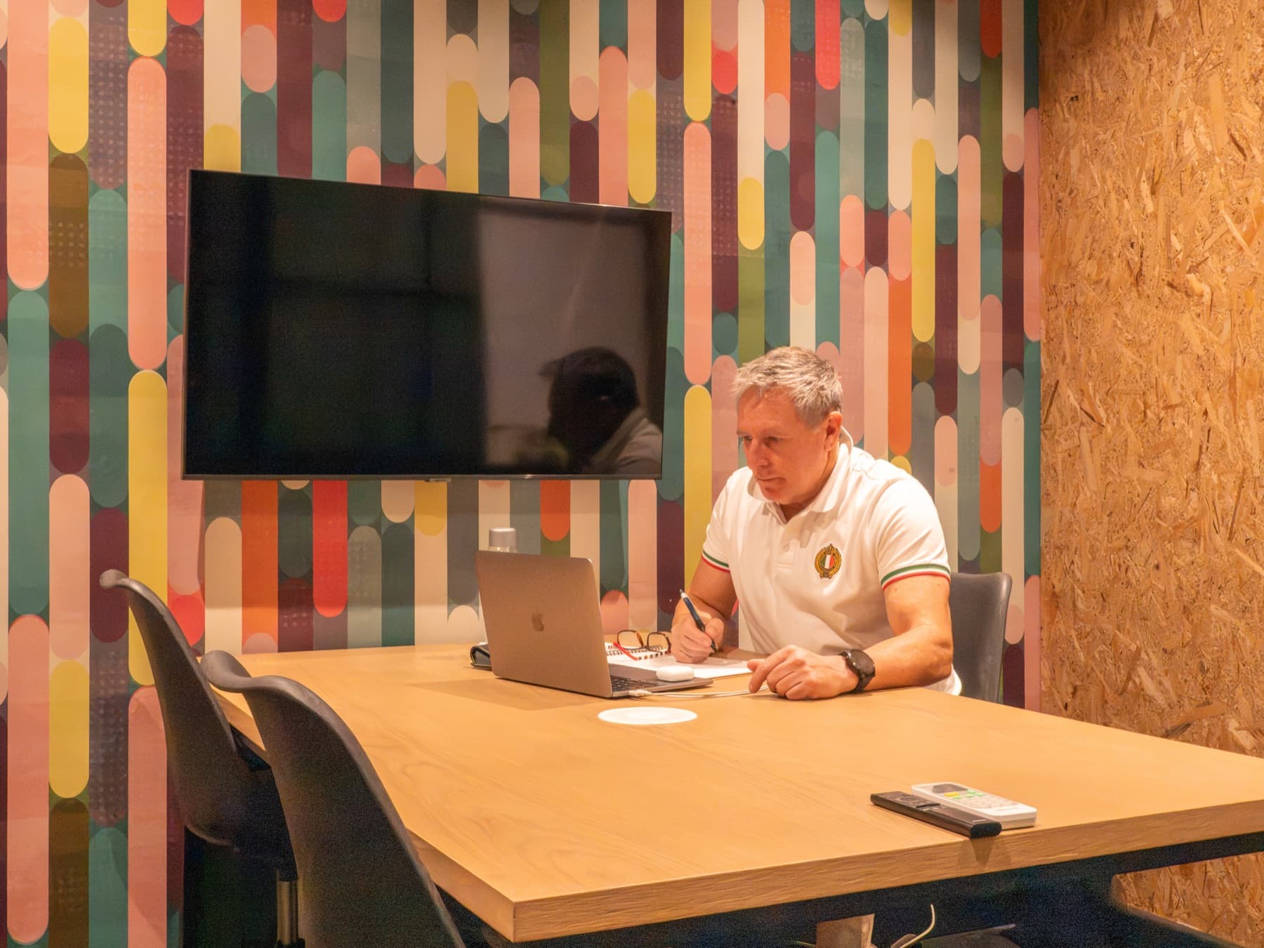 A man at the table in a SOHO meeting room, beneath a wall-mounted presentation screen.